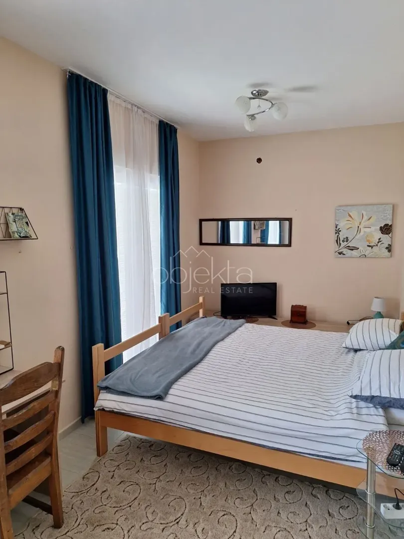 Namještena spavaća soba sa bračnim krevetom, prozorom sa plavim zavjesama i televizorom.