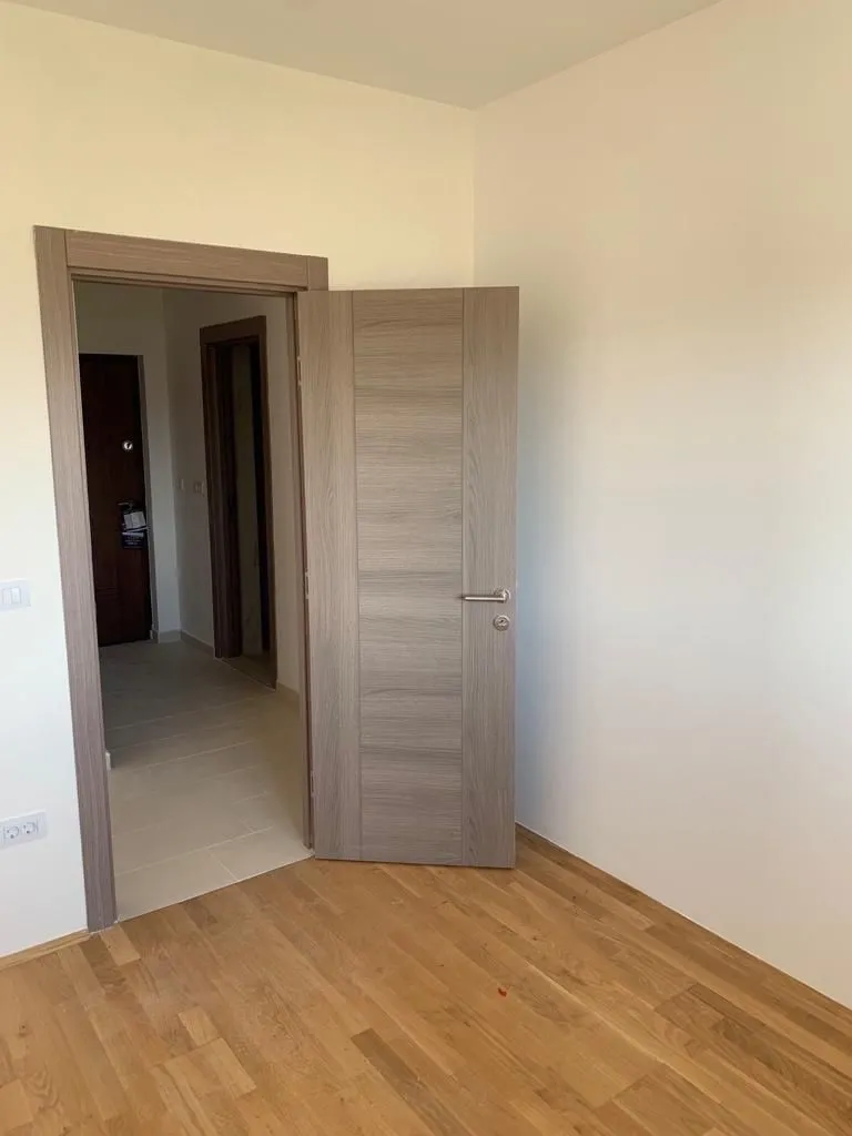 Prazna soba sa drvenim podom i otvorenim vratima koja vode u hodnik.
