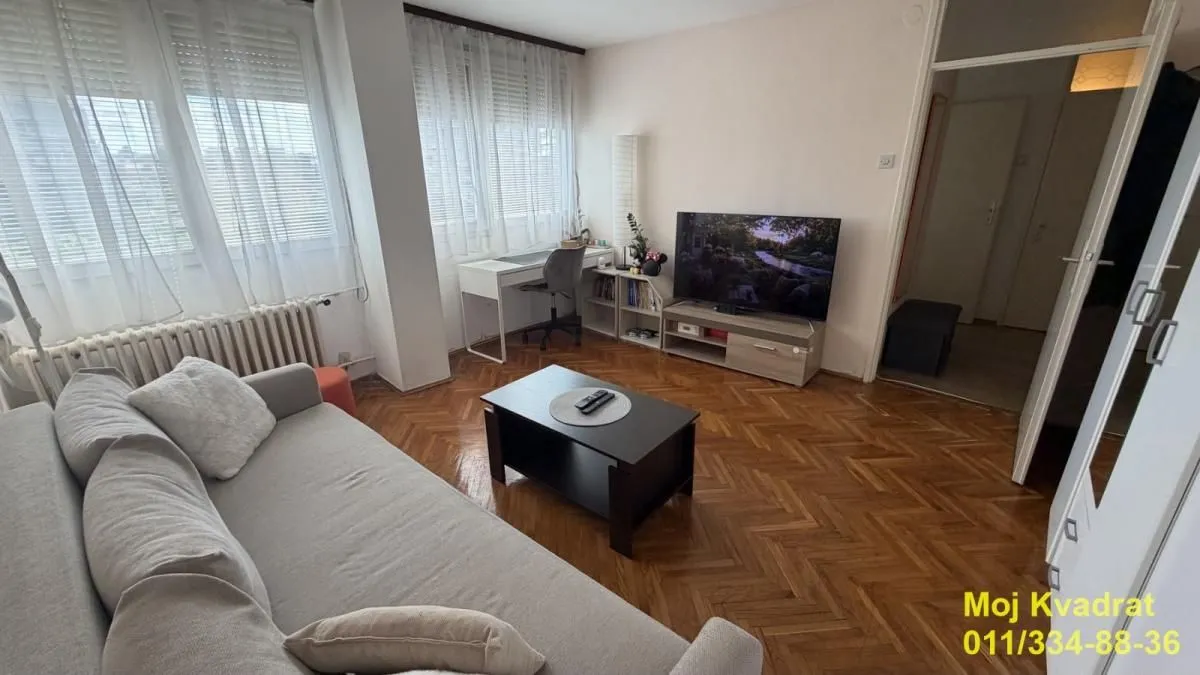 Dnevna soba sa garniturom, televizorom i radnim stolom pored prozora.