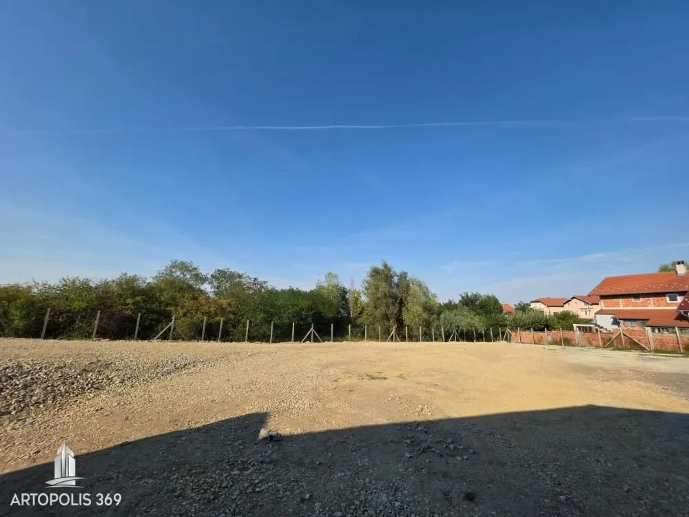 Pogled na ograđeni plac sa rastinjem i okolnim kućama u pozadini.