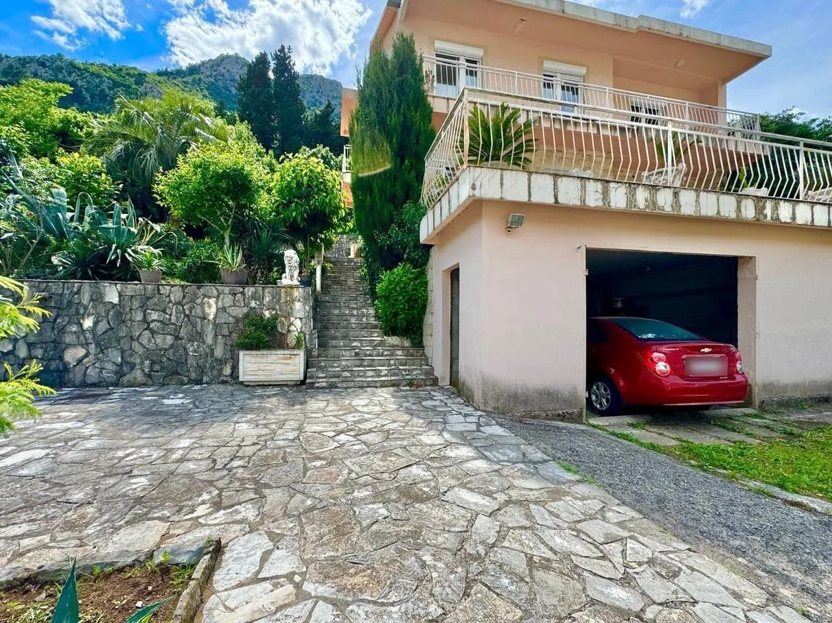 Dvorište kuće sa popločanim prilazom, garažom, terasom i stepeništem koje vodi u zelenilo.