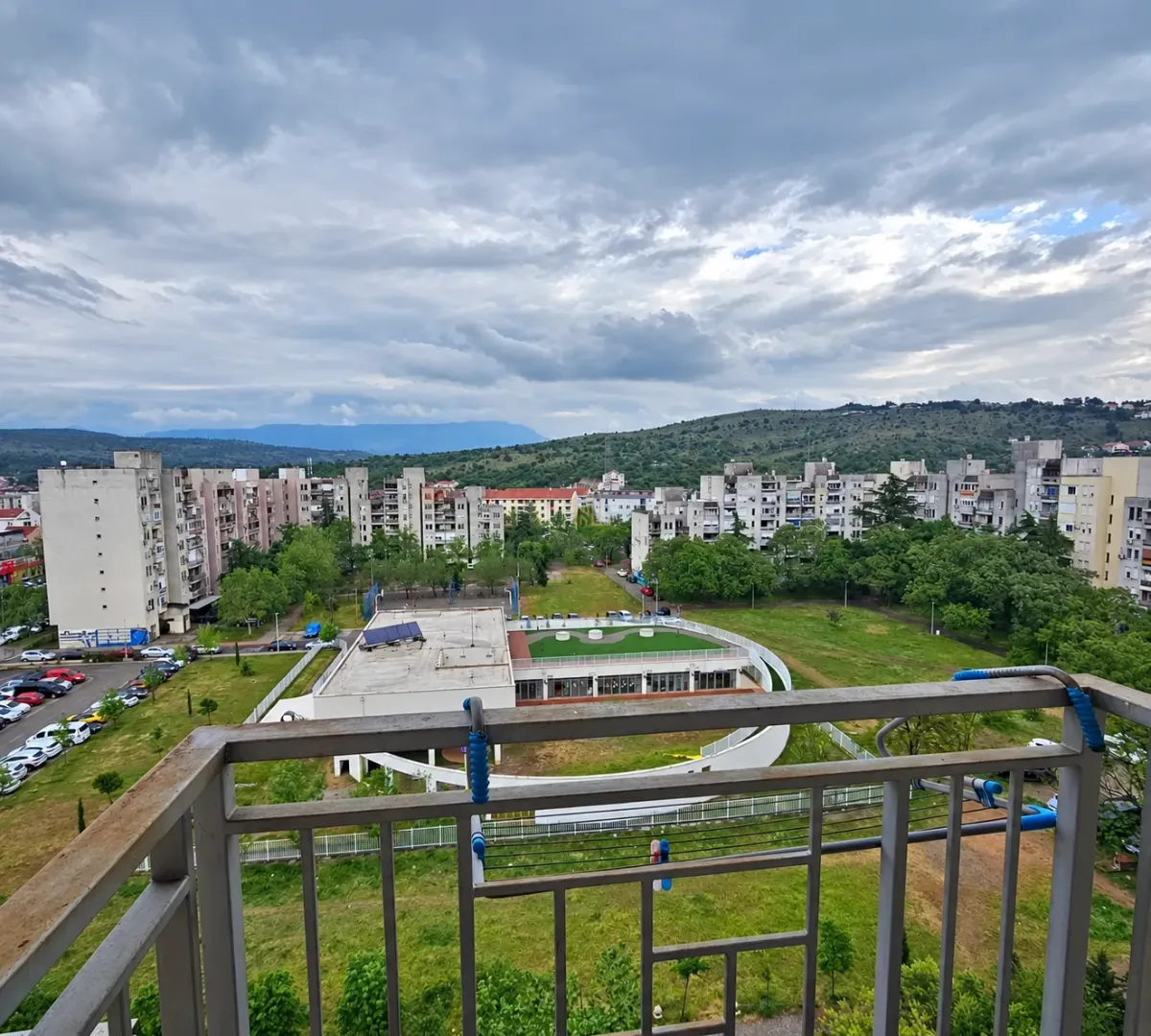 Pogled sa terase na okolne stambene zgrade, zelenilo i brda u daljini.