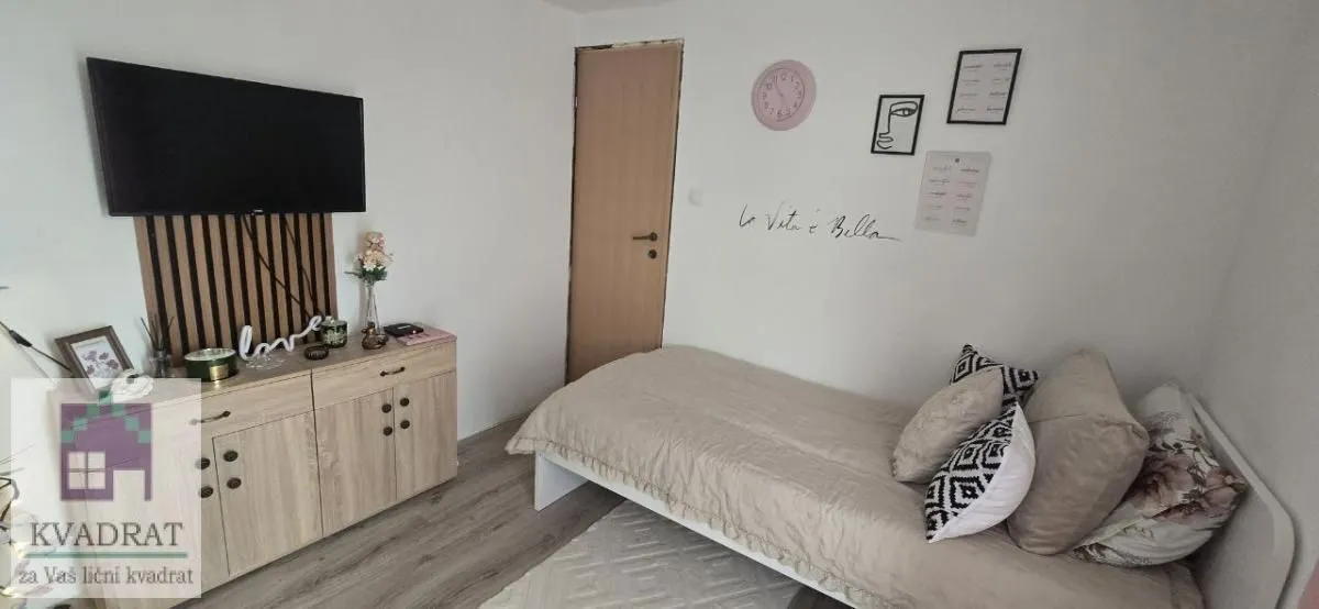 Manja spavaća soba sa krevetom, komodom i televizorom na zidu.