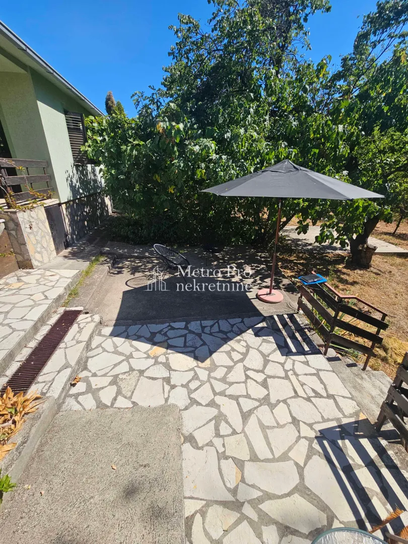 Popločano dvorište kuće sa garniturom za sedenje i zelenilom.