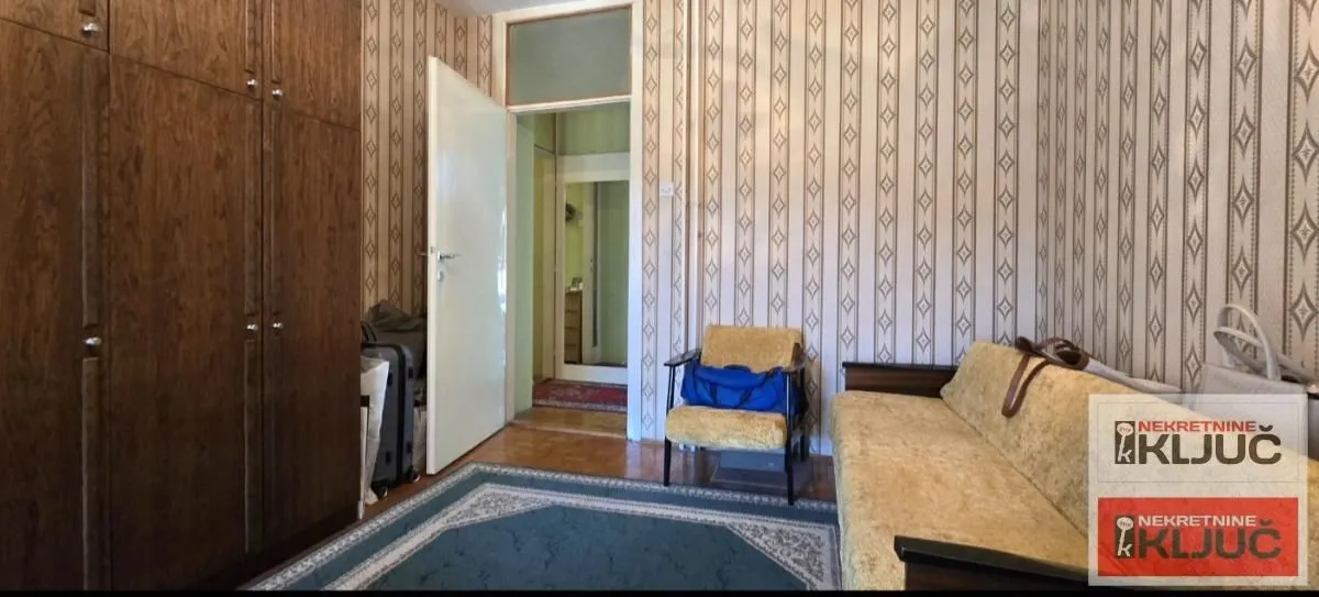 Dnevna soba sa garniturom, velikim drvenim ormarom i tapetama na zidovima.