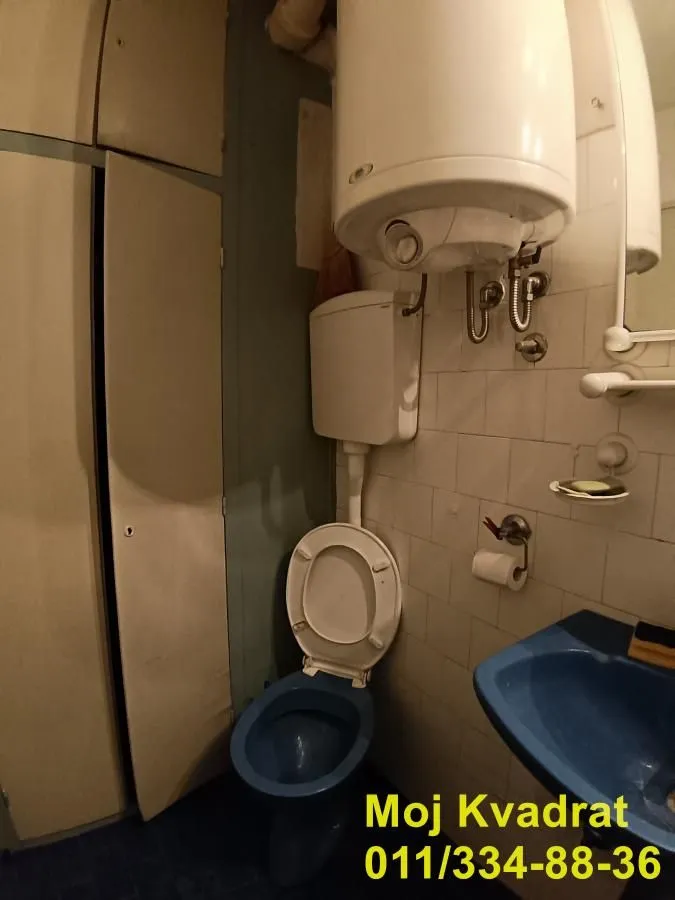 Manje kupatilo sa plavom wc šoljom i umivaonikom, te bojlerom na zidu.