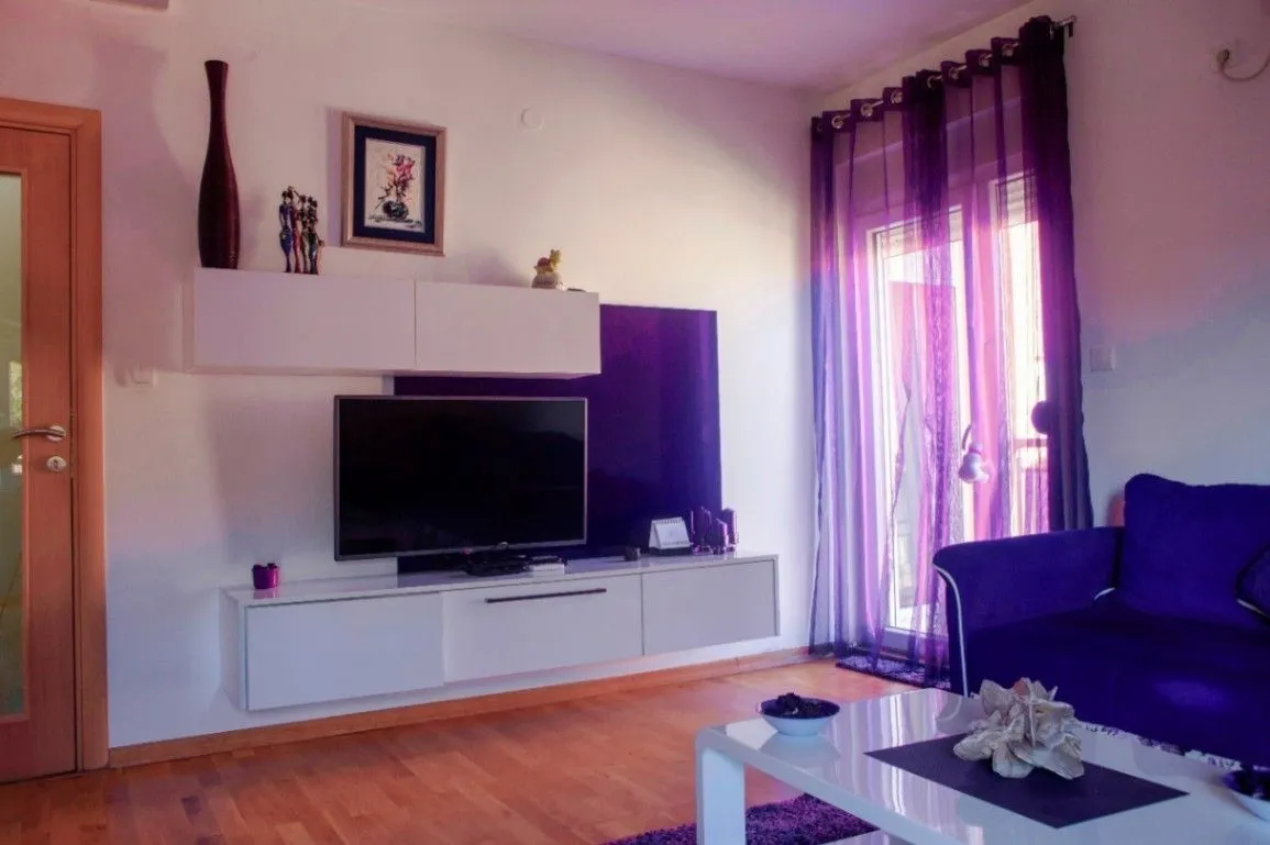 Svijetla dnevna soba sa ljubičastim detaljima, garniturom i televizorom.