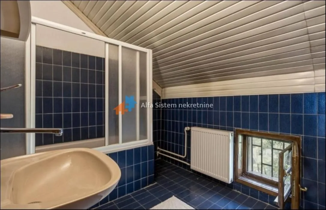 Kupatilo sa plavim pločicama, umivaonikom i tuš kabinom u potkrovlju.