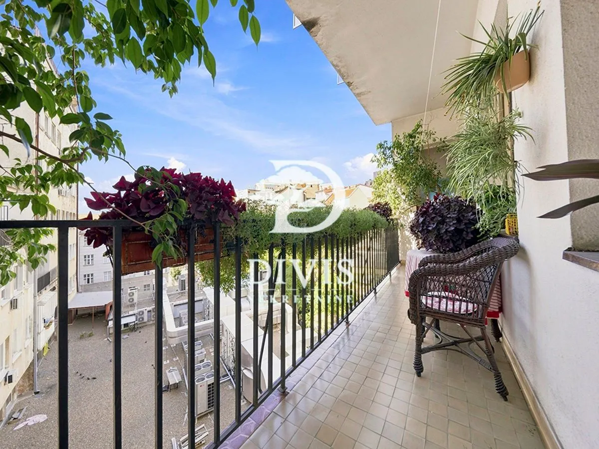 Natkriveni balkon sa metalnom ogradom, ukrašen saksijama sa cvećem i pruža pogled na grad.