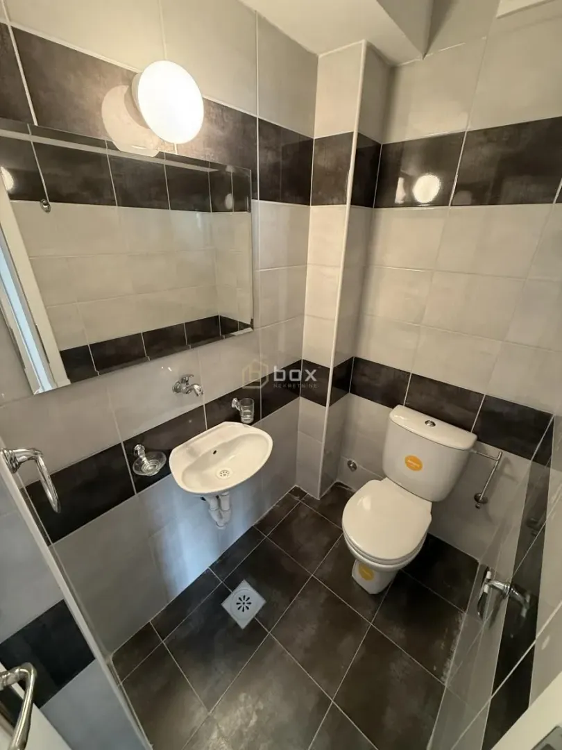 Small bathroom with a sink, mirror, and toilet, tiled with grey tiles.