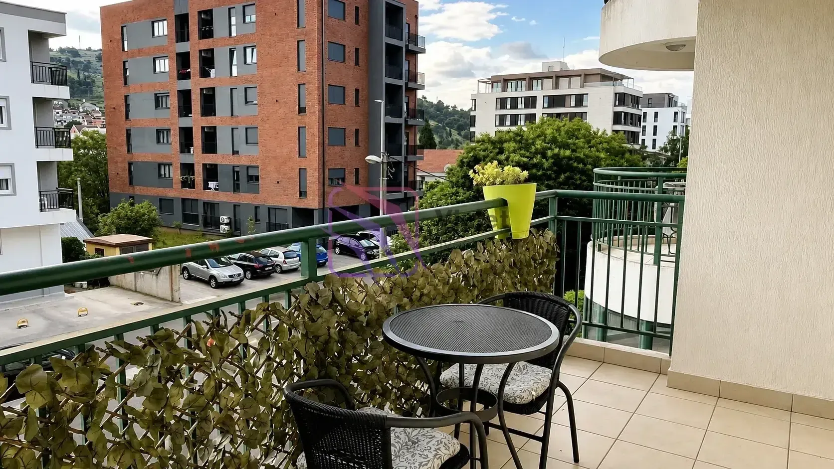 Namješten balkon sa stolom i stolicama i pogledom na okolne zgrade i parking.