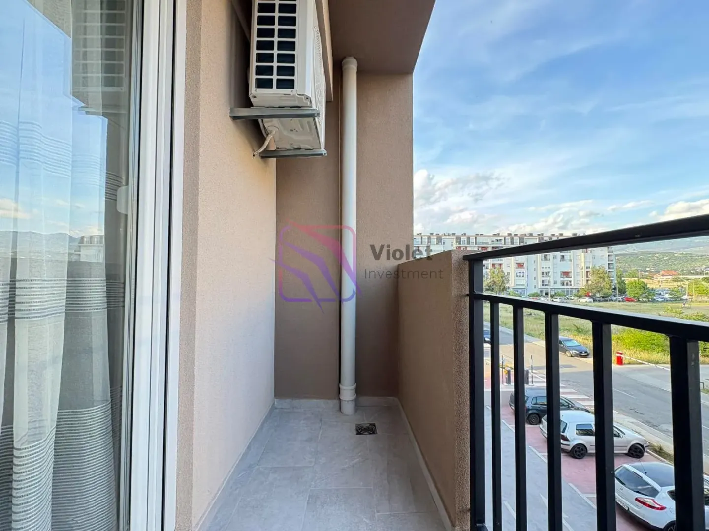 View from the tiled balcony with an air conditioning unit and railing overlooking surrounding buildings and parking.