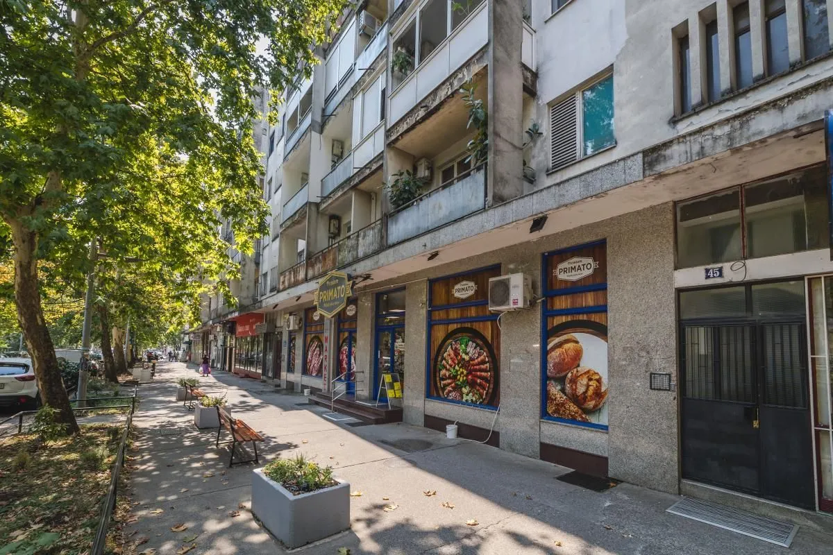 Pogled na spoljašnjost zgrade sa trotoarom i lokalom u prizemlju.