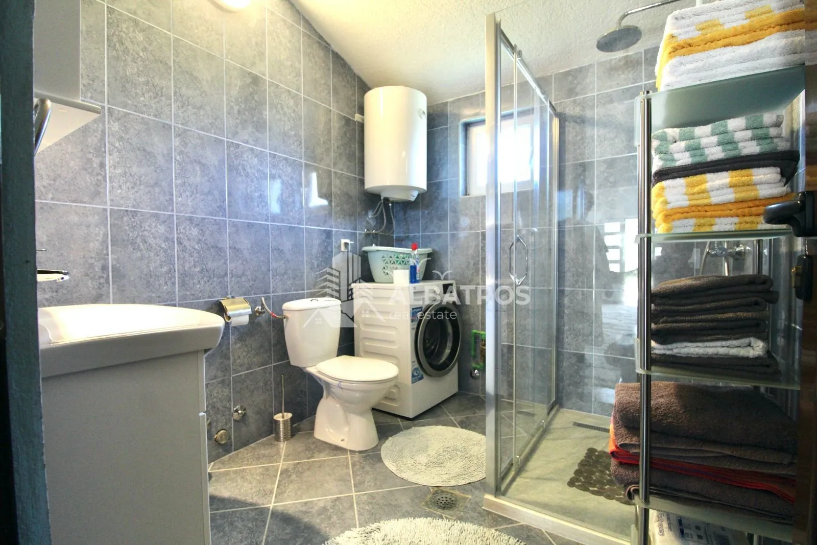 Kupatilo obloženo sivim pločicama sa tuš kabinom, WC šoljom, umivaonikom i veš mašinom.