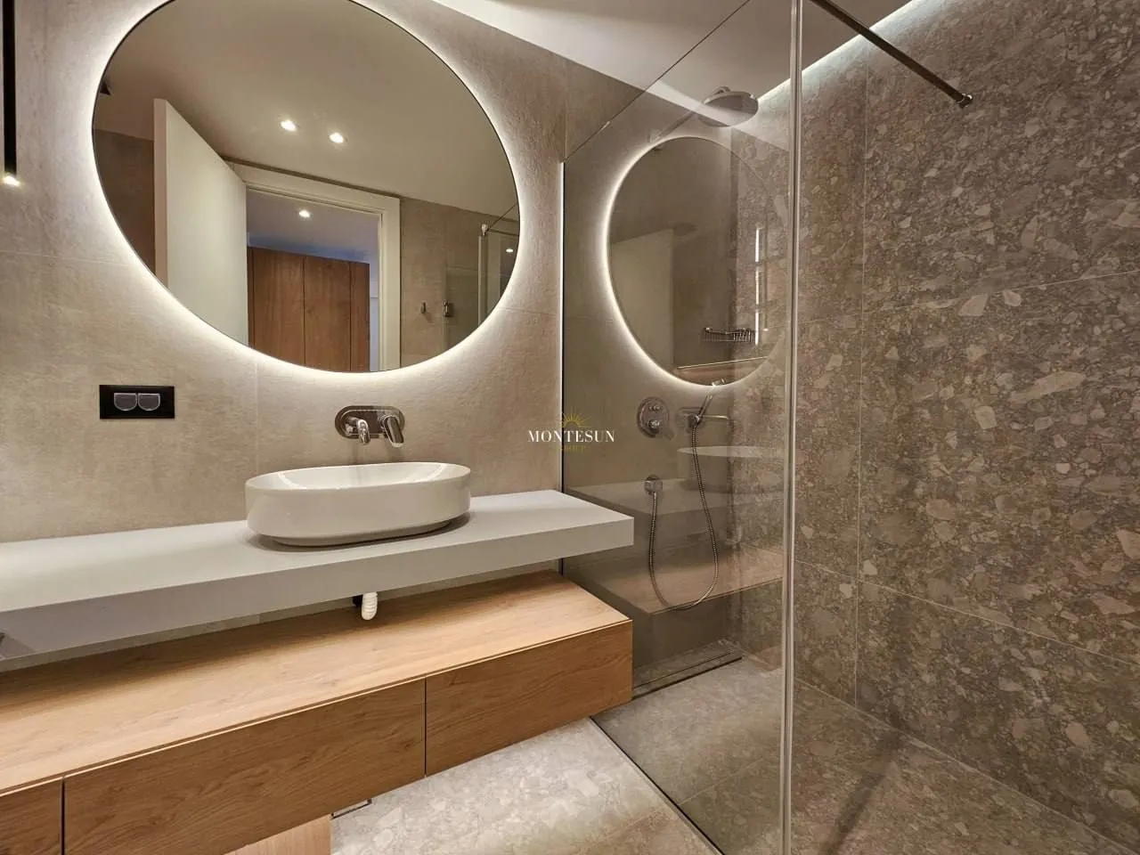 Modern bathroom with a sink, large round illuminated mirror, and a walk-in shower.