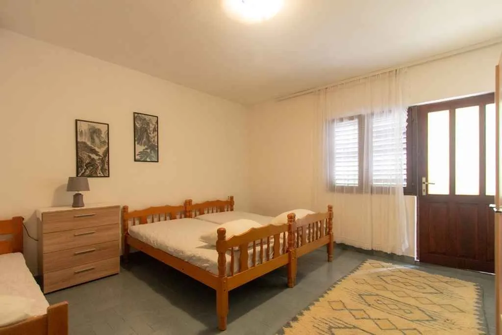 Spavaća soba sa drvenim krevetima, komodom i izlazom na terasu.