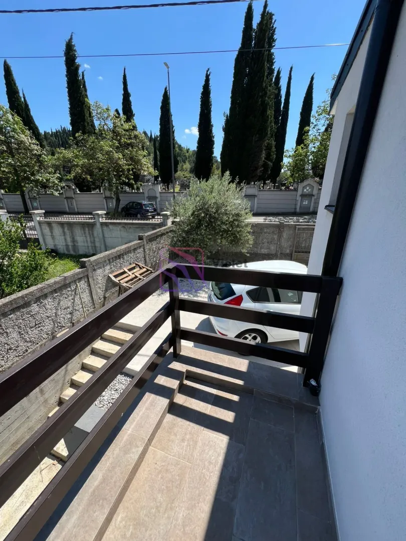 Manji popločani balkon sa tamnom drvenom ogradom i pogledom na dvorište i okolinu.