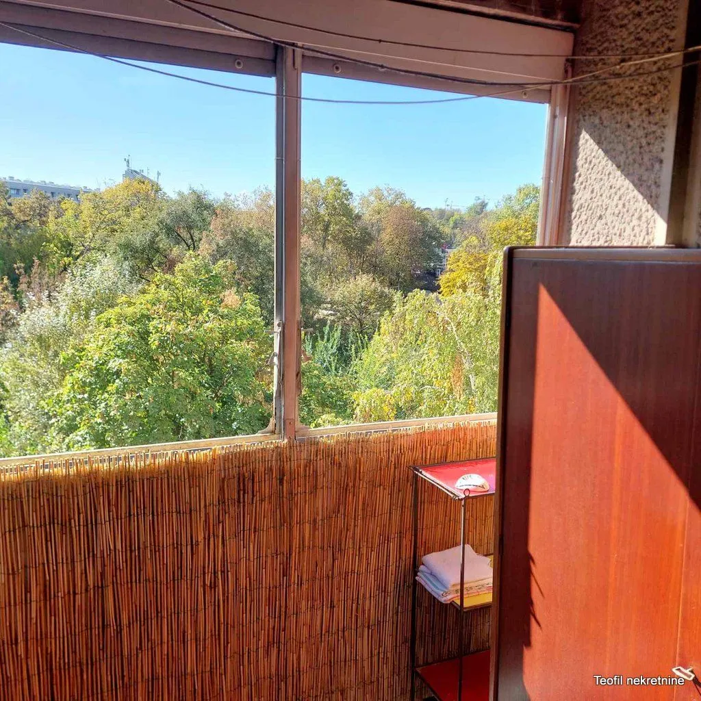 Manji zatvoreni balkon sa pogledom na zelene krošnje drveća.