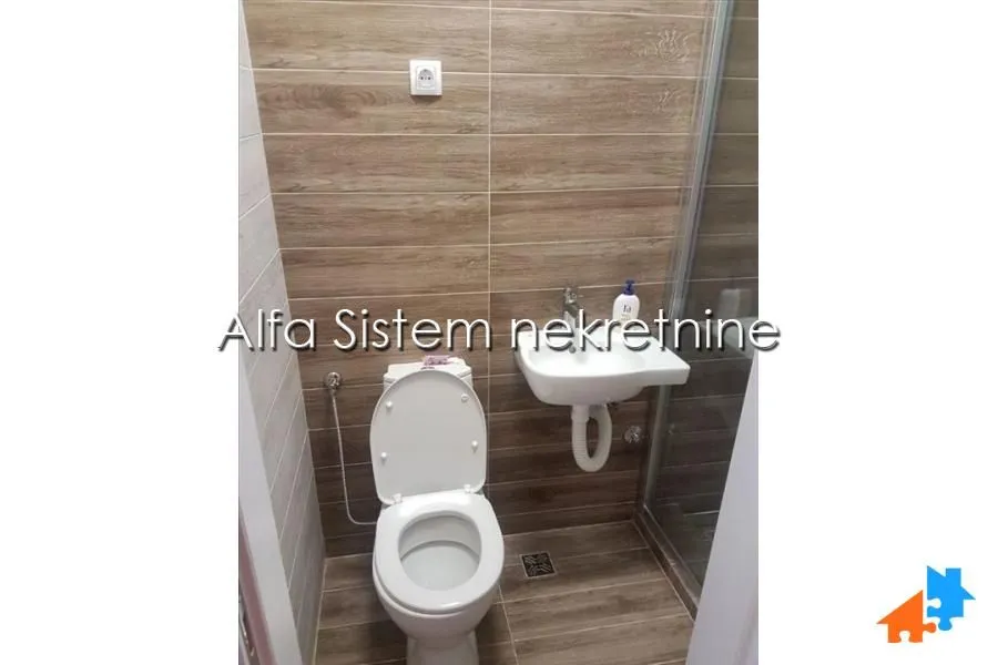 Manje kupatilo sa WC šoljom, umivaonikom i tuš kabinom obloženo pločicama u dezenu drveta.