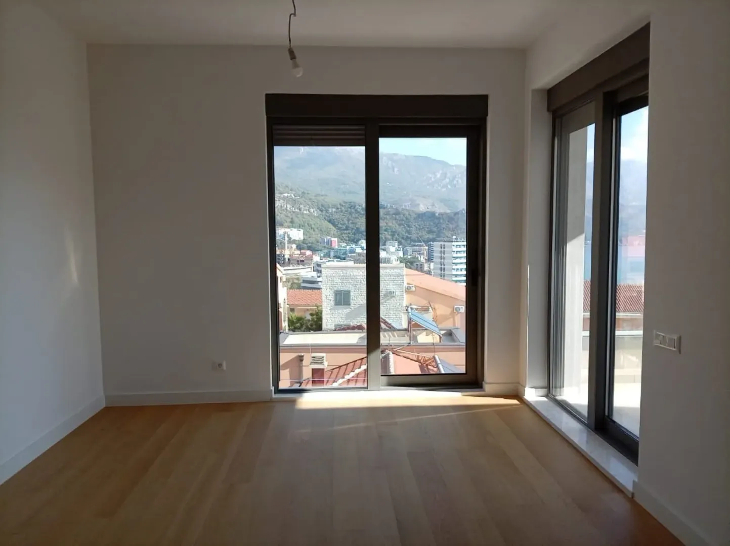Prazna soba sa parketom, velikim staklenim površinama i pogledom na grad i planine.