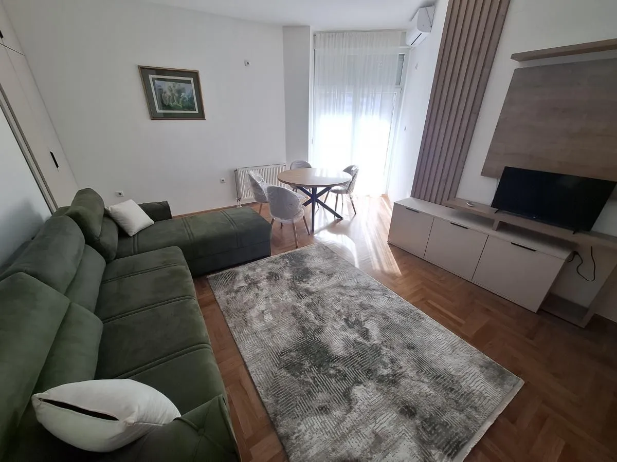 Dnevna soba sa ugaonom garniturom, televizorom i trpezarijskim stolom kod prozora.