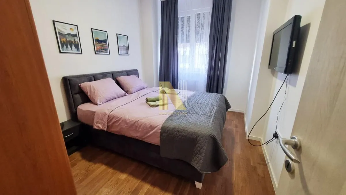 Nameštena spavaća soba sa bračnim krevetom, TV-om i prozorom sa zavesama.