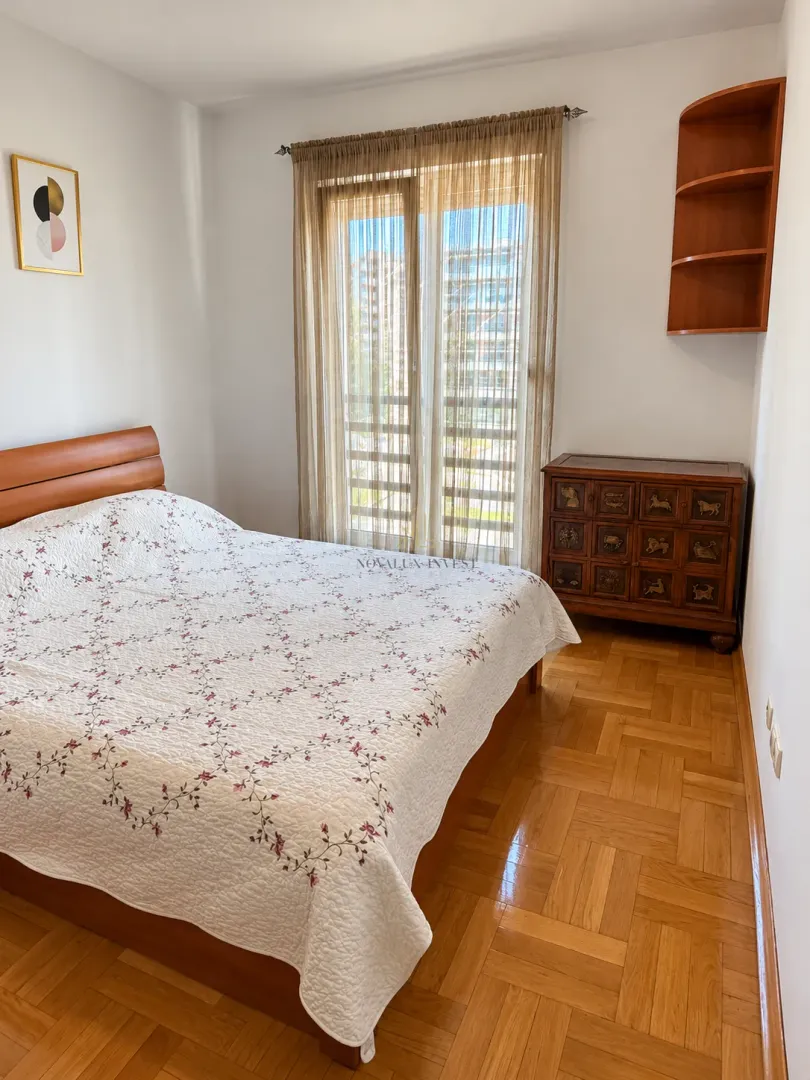 Namještena spavaća soba sa bračnim krevetom, drvenom komodom i izlazom na balkon.