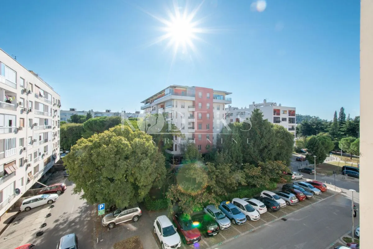 Pogled sa prozora na okolne zgrade, parking i zelene površine.