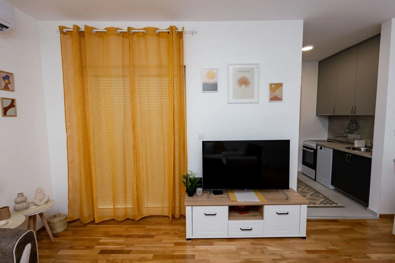 Dnevna soba sa televizorom, žutim zavesama i pogledom na kuhinju.