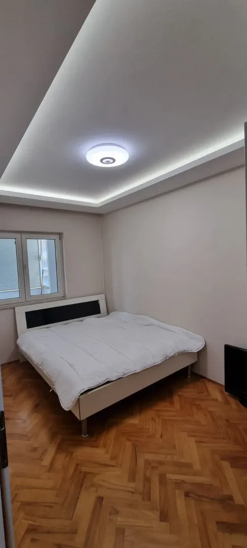 Svijetla spavaća soba sa bračnim krevetom, parketom i dekorativnom rasvjetom na plafonu.