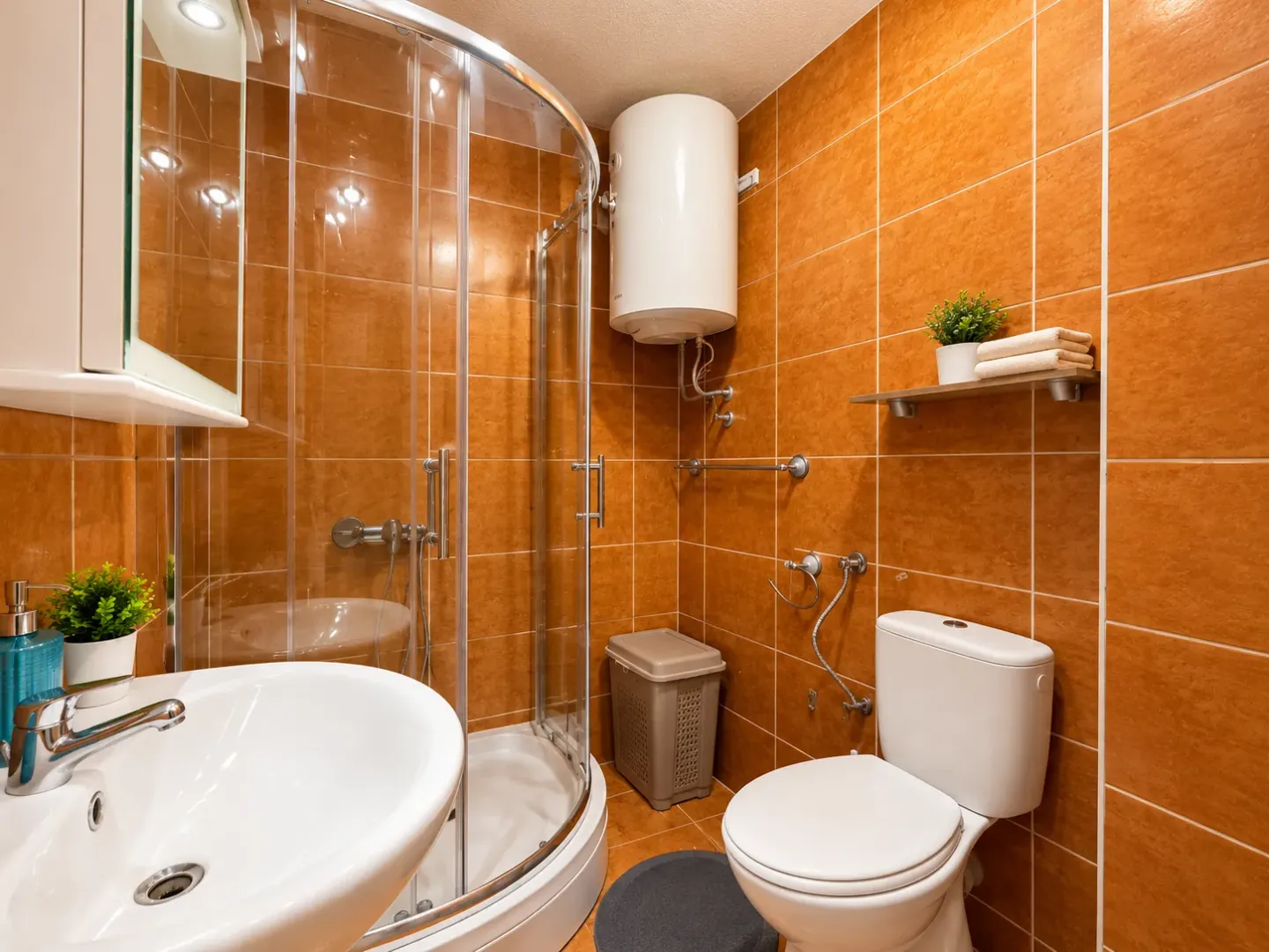 Moderne kupatilo sa tuš kabinom, umivaonikom i WC šoljom obloženo narandžastim pločicama.