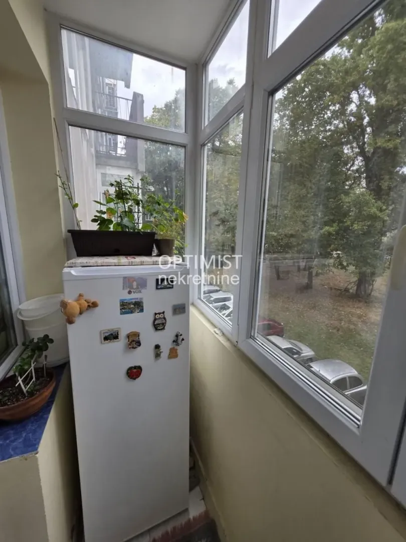 Zastakljeni balkon sa frižiderom i pogledom na zelenilo.