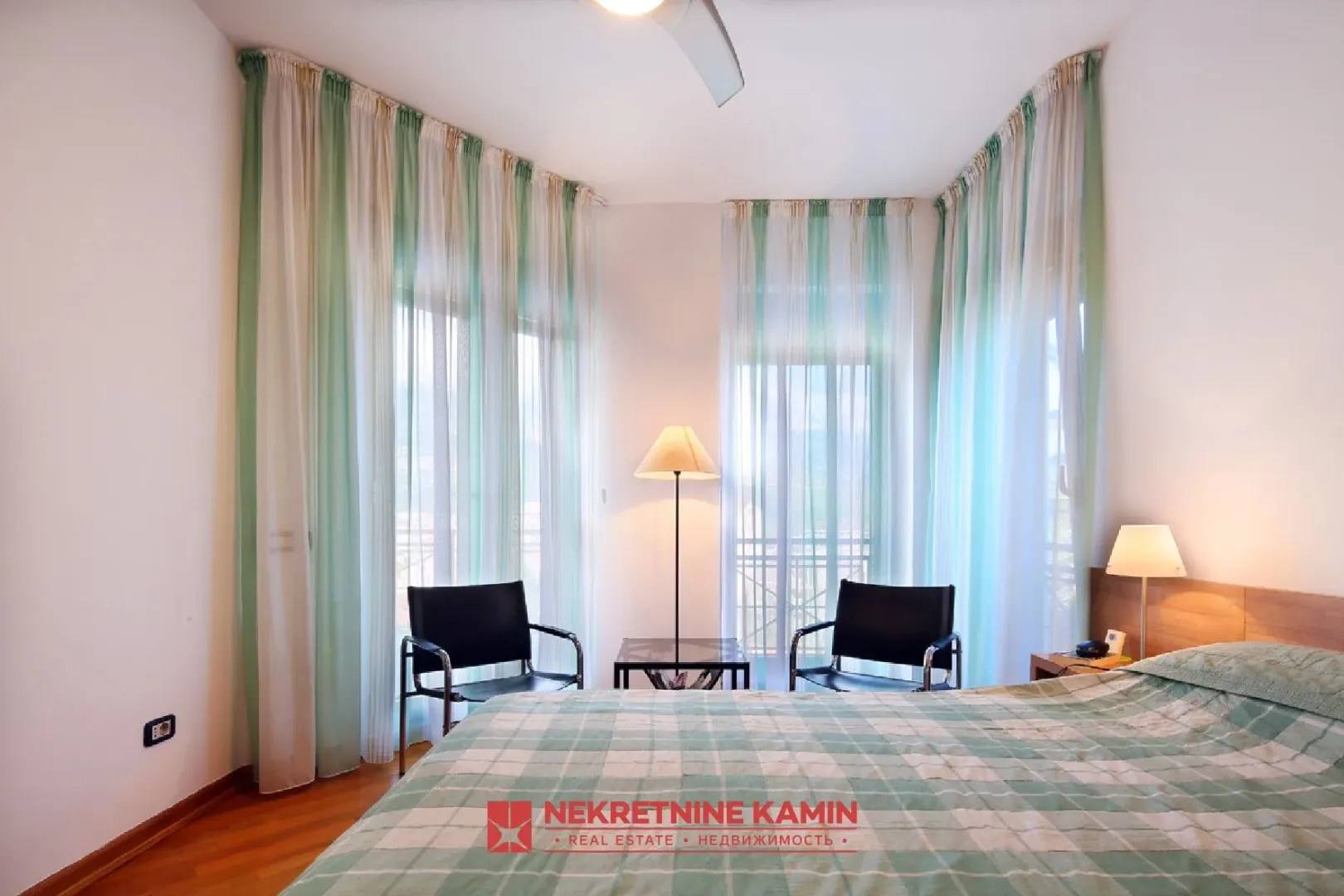 Furnished bedroom with a double bed, armchairs, and large windows with light curtains.