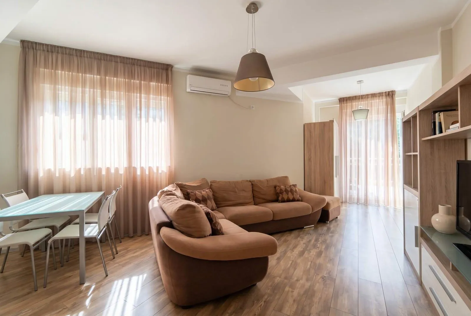 Svijetla dnevna soba sa ugaonom garniturom, trpezarijskim stolom i modernim osvjetljenjem.