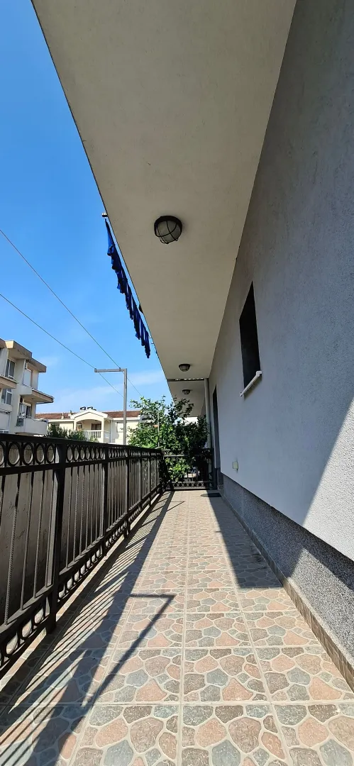 Dugački popločani balkon sa metalnom ogradom i pogledom na okolne zgrade.