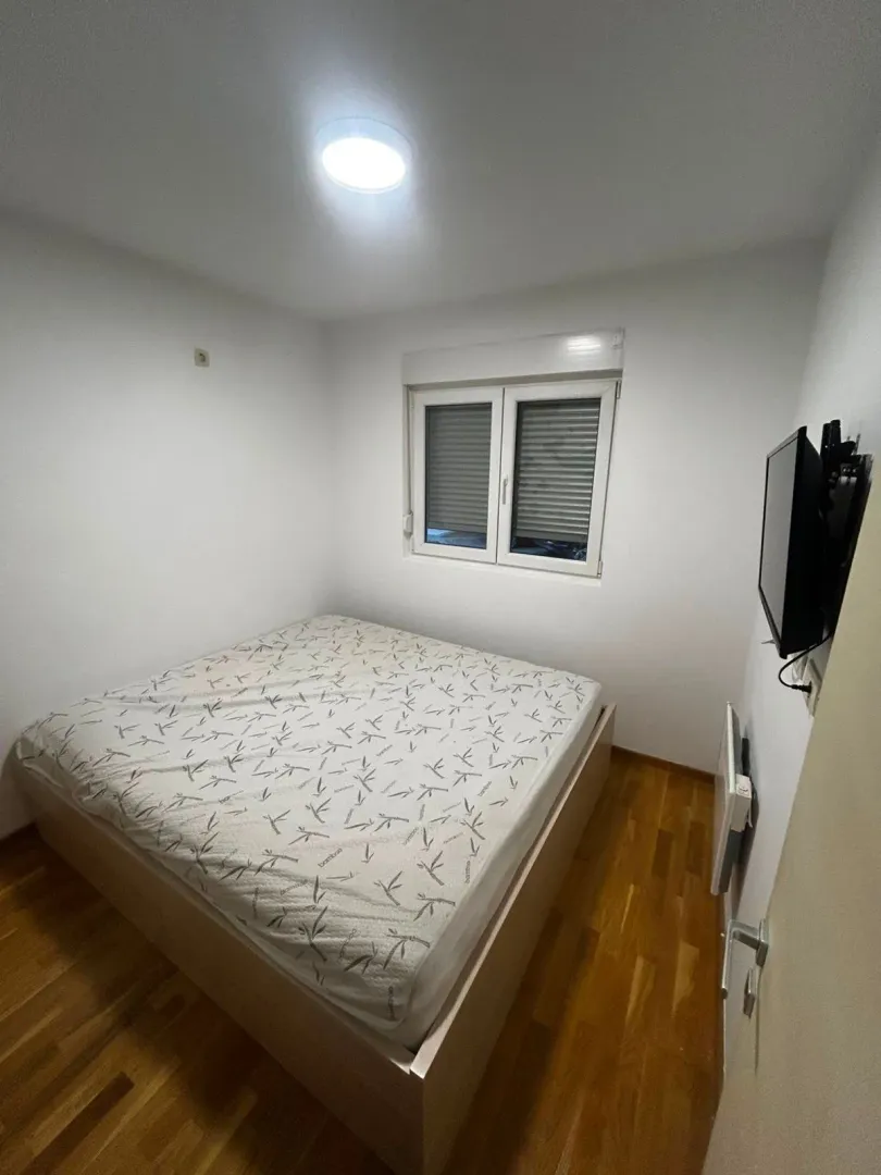 Manja spavaća soba sa bračnim krevetom, drvenim podom i prozorom.