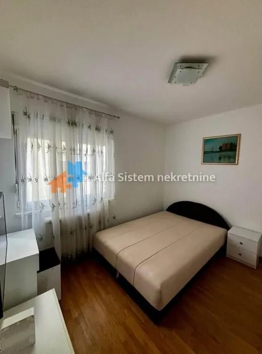 Nameštena spavaća soba sa bračnim krevetom, prozorom sa zavesama i noćnim stočićem.