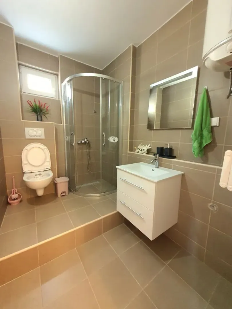Uređeno kupatilo sa tuš kabinom, umivaonikom, ogledalom i WC šoljom u bež tonovima.