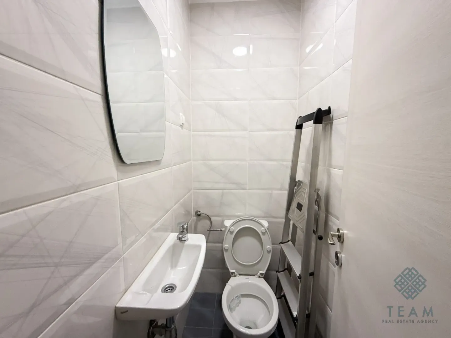 Manje kupatilo sa bijelim pločicama, umivaonikom, WC šoljom i ogledalom.