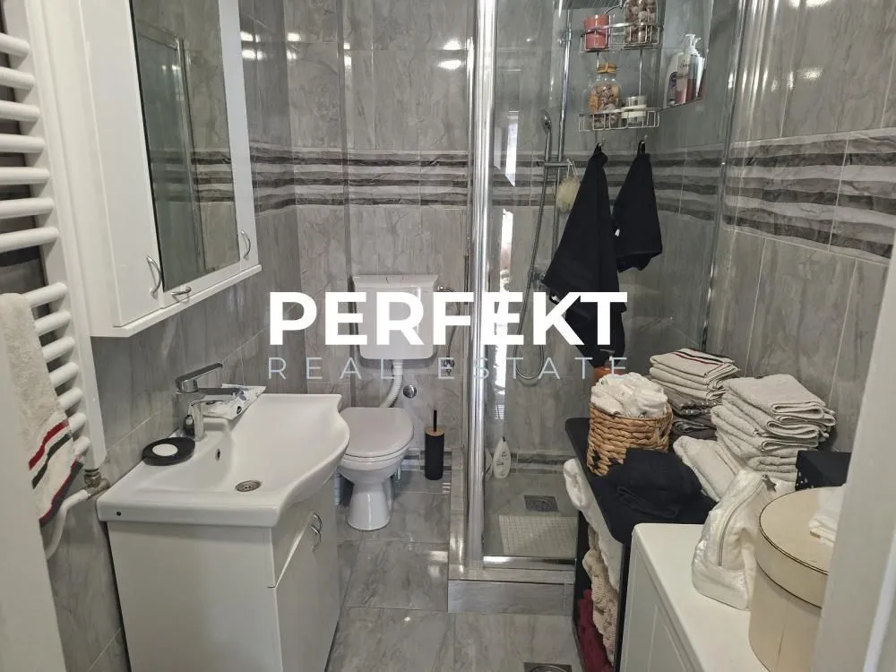 Kompaktno kupatilo sa tuš kabinom, umivaonikom i WC šoljom obloženo sivim pločicama.