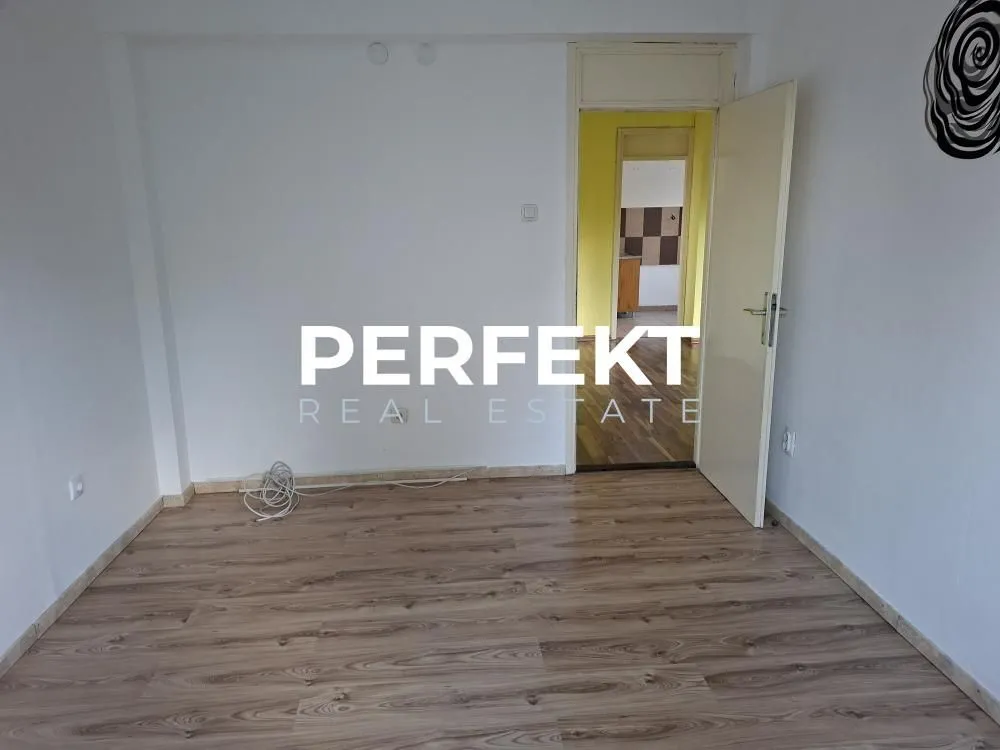 Prazna soba sa laminatom i otvorenim vratima prema hodniku.