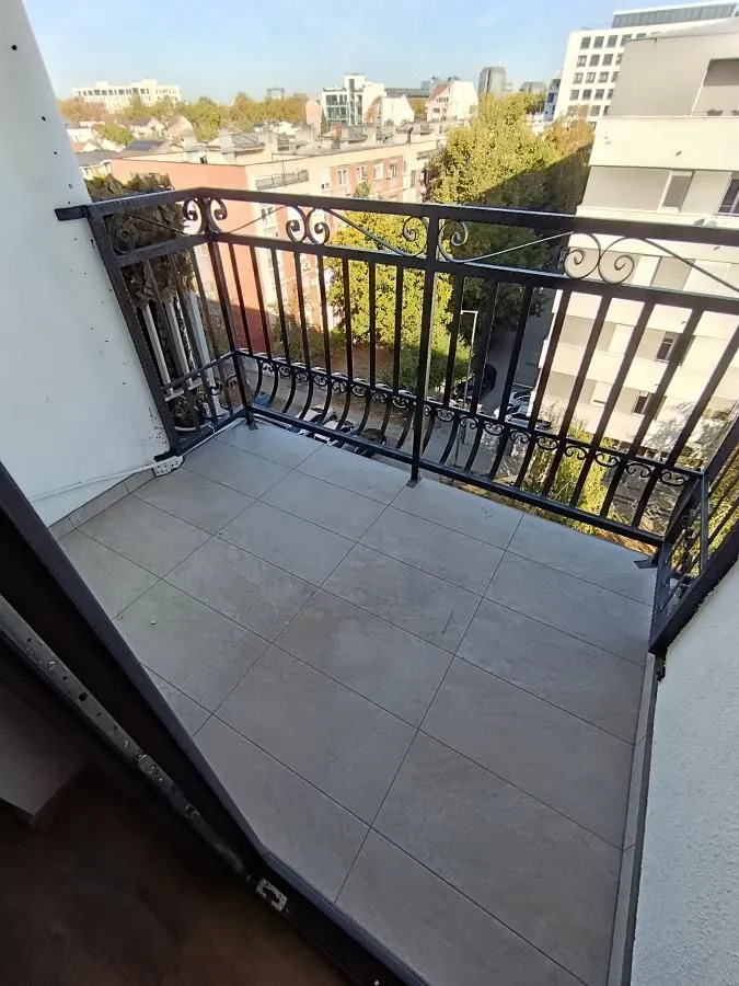 Manji popločani balkon sa ukrasnom metalnom ogradom i pogledom na okolne zgrade.