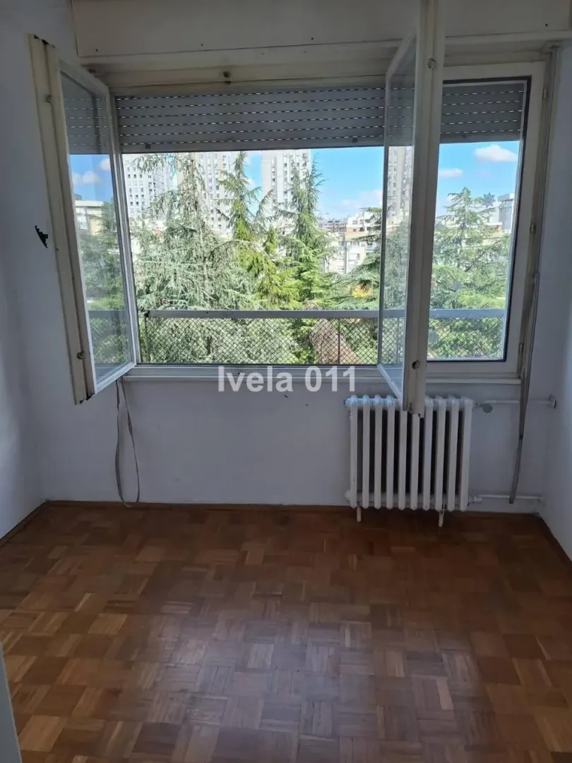 Empty room with parquet flooring and an open window with a view of greenery and buildings.