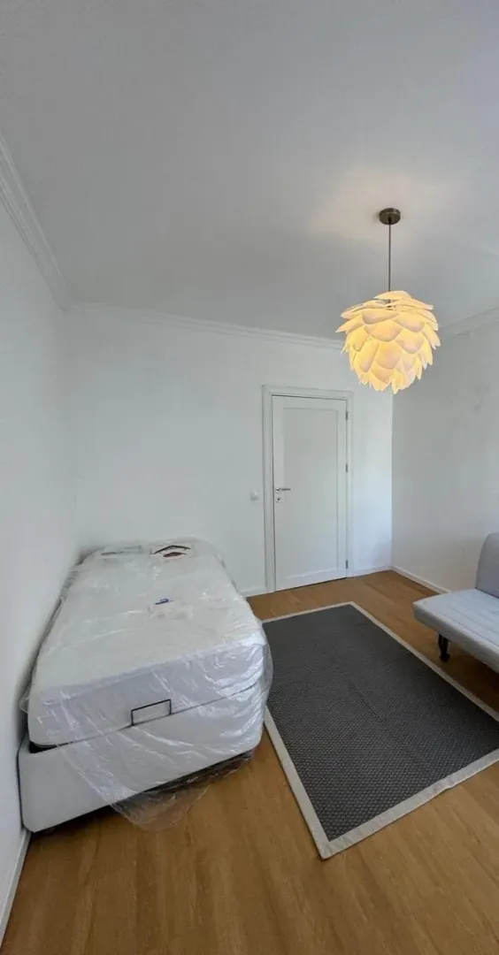 Small bedroom with a bed covered in protective film and a rug.