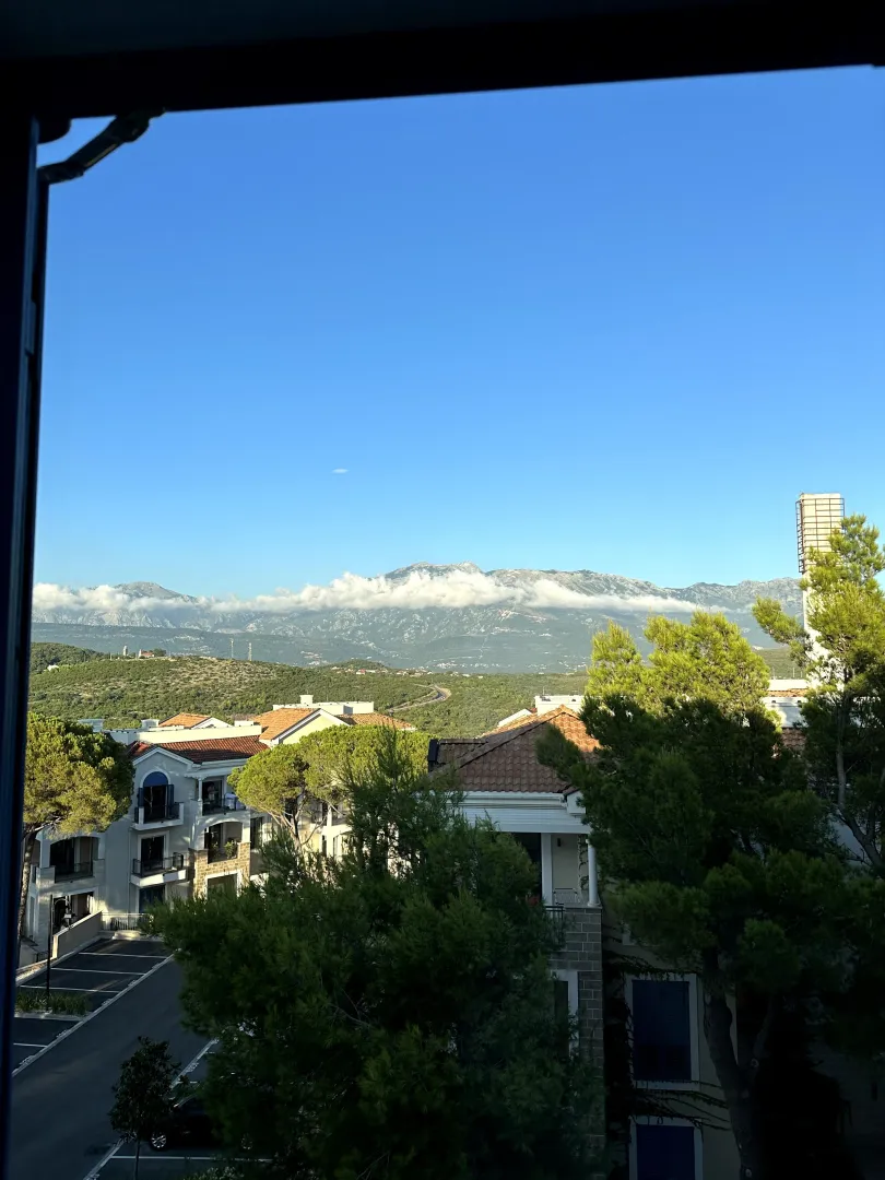 Pogled sa prozora na okolne zgrade, zelenilo i planine u pozadini.