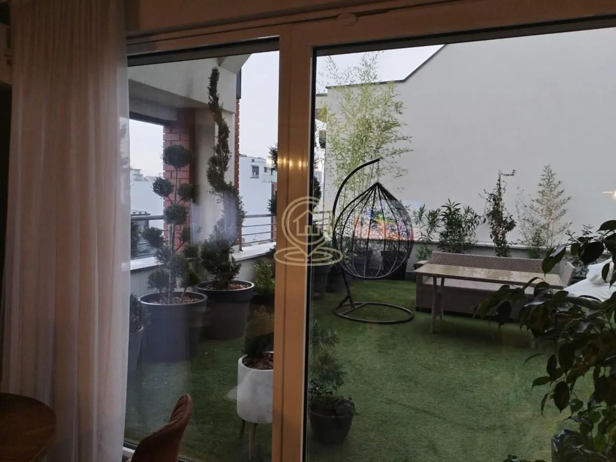 Pogled kroz balkonska vrata na uređenu terasu sa veštačkom travom i garniturom za sedenje.