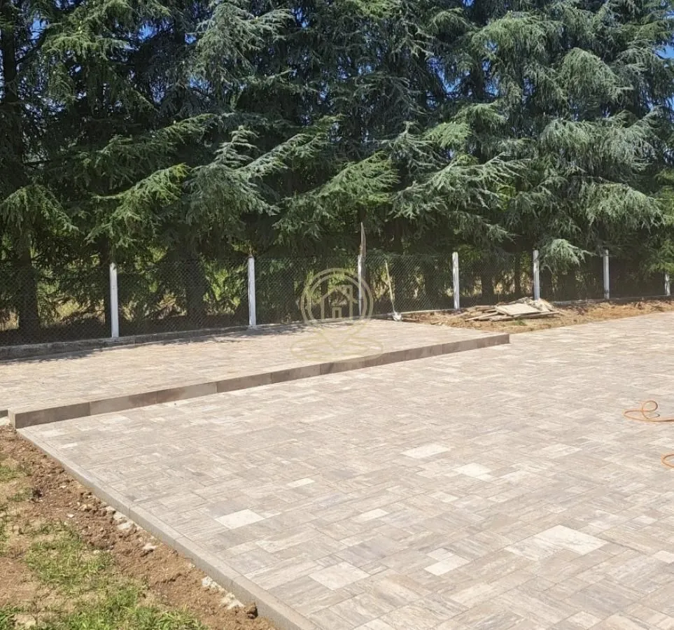 Popločano dvorište sa ogradom i zelenilom u pozadini.