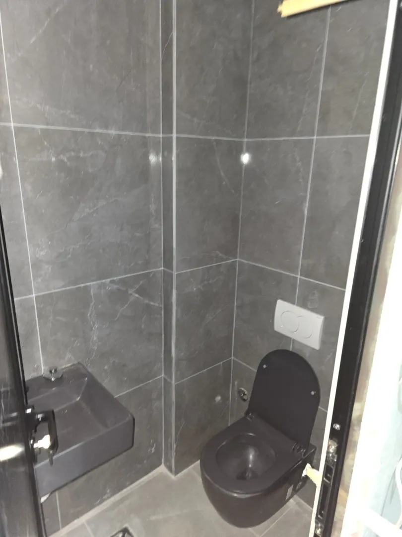 Small toilet room with grey tiles, a dark toilet, and a small sink.