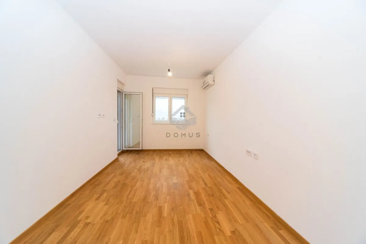 Prazna soba sa bijelim zidovima, drvenim parketom i izlazom na terasu.