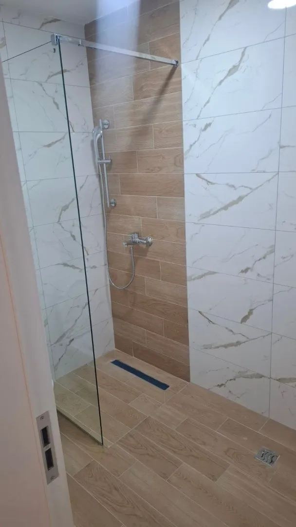 Moderno opremljena tuš kabina sa staklenom pregradom i kombinacijom pločica u dezenu drveta i mermera.